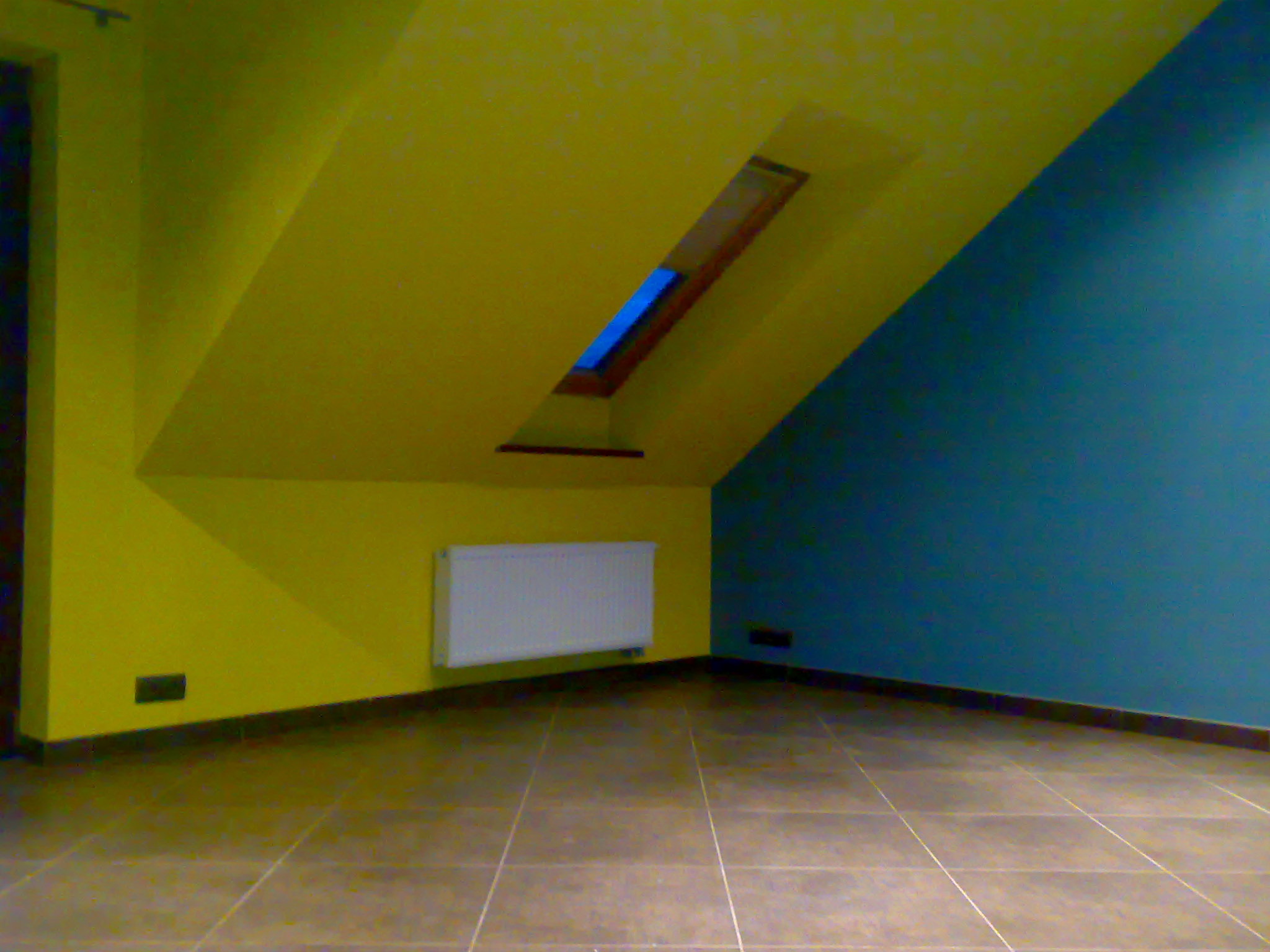 Poddasze po remoncie: żółta ściana z oknem dachowym i grzejnikiem, niebieska ściana boczna, brązowe płytki na podłodze.